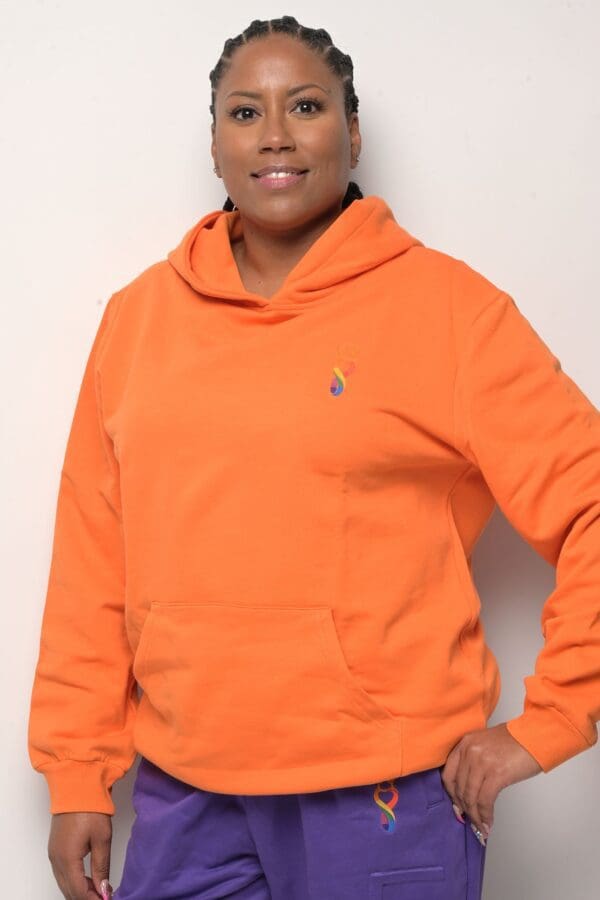 A woman in an orange hoodie standing next to a wall.