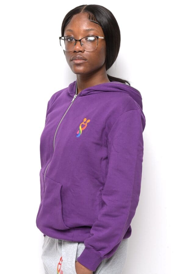 A woman in purple hoodie standing against a white wall.